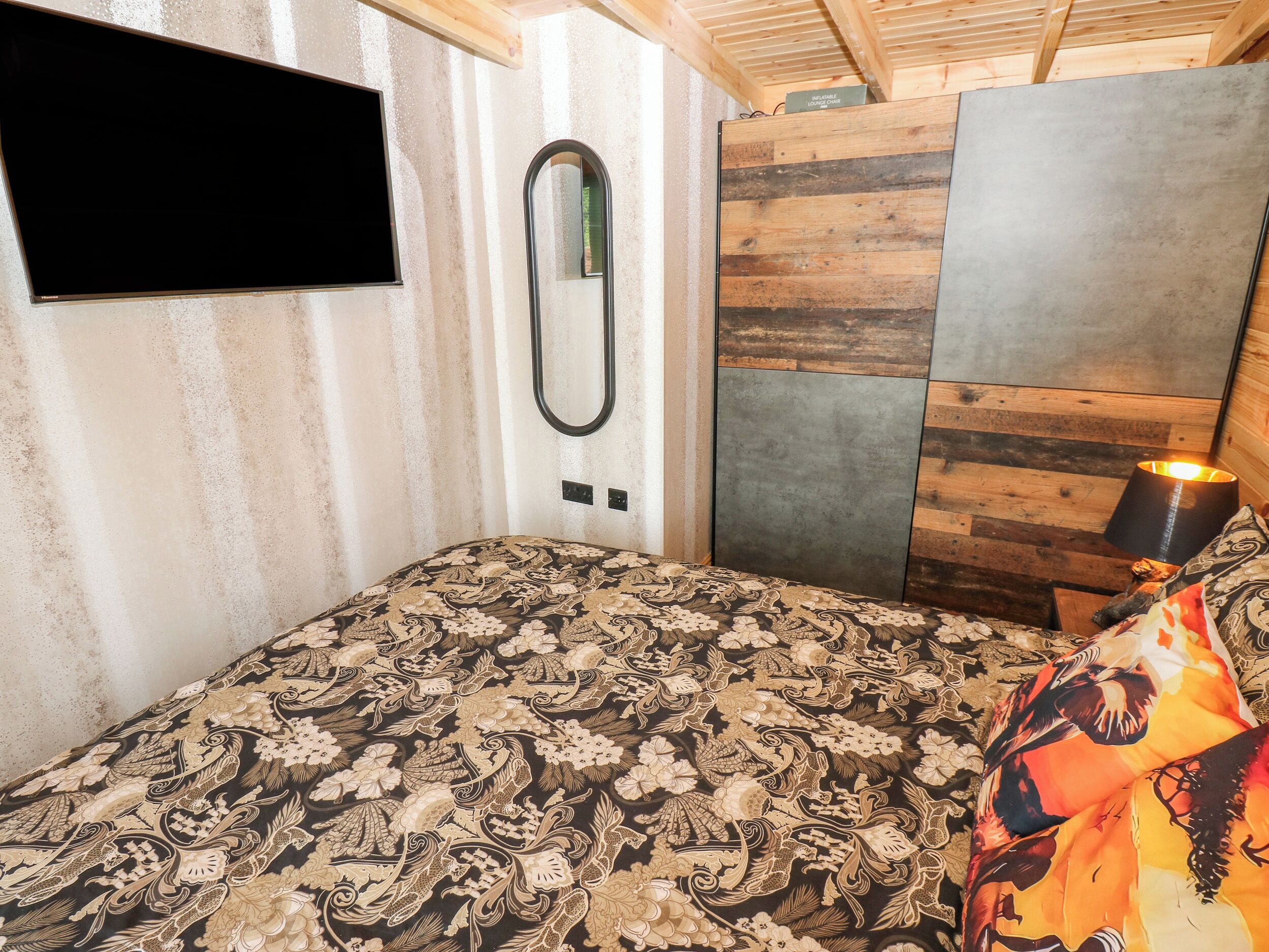 A bedroom with a patterned bedspread, a mounted mirror, a flat-screen TV, a wooden headboard, and a bedside lamp.