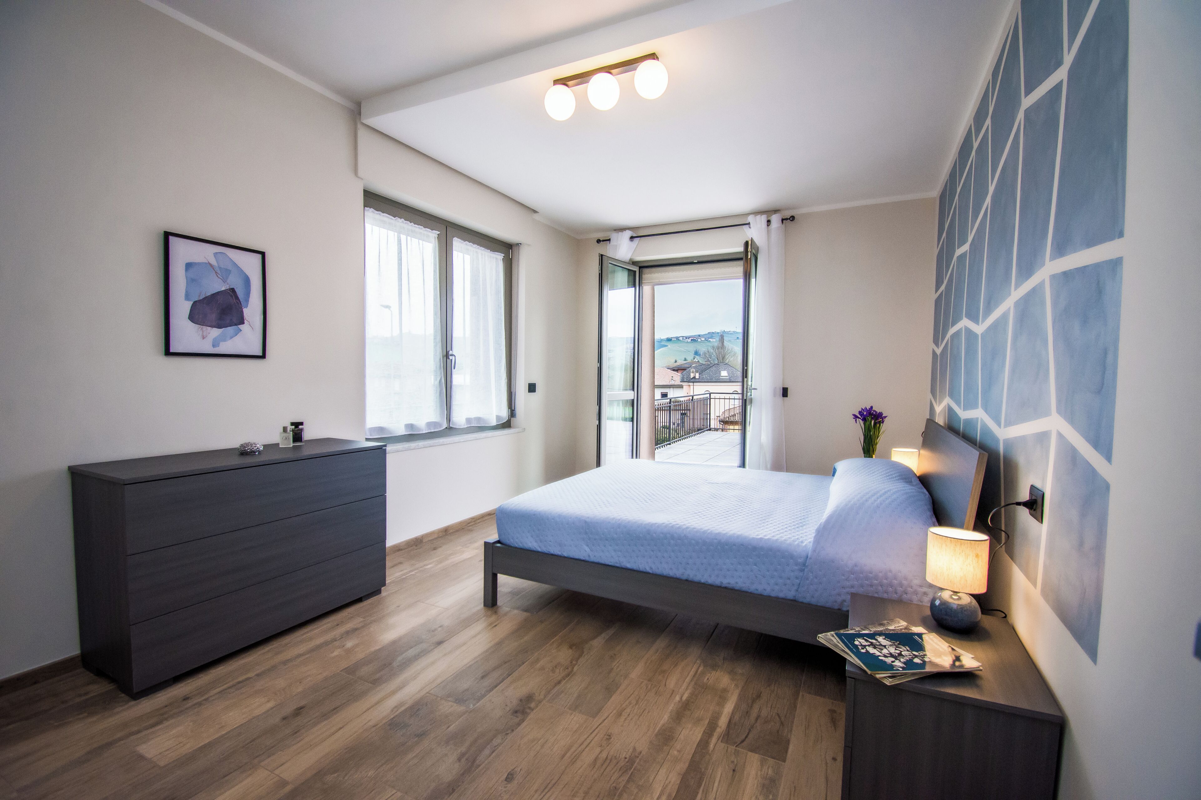 A modern bedroom with a bed, dresser, and a view through a large window.