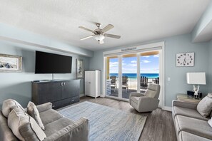 A living room with a flat-screen TV, a ceiling fan, a large window with a view of the beach, and a sliding glass door leading to an outdoor seating area. - stunning Gulf views, resort-style amenities, and an unbeatable location (Panama City Beach)