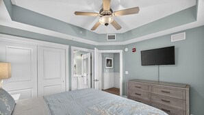 Un dormitorio con cama, cómoda, ventilador de techo, televisor y puerta que da a otra habitación.