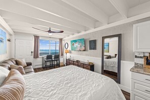 Une chambre avec un grand lit, un bureau, une chaise, un ventilateur de plafond, une fenêtre avec des rideaux et une vue sur l’océan.