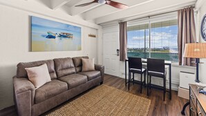 Ein Wohnzimmer mit einem braunen Ledersofa, einem Esstisch mit Stühlen, einem Deckenventilator, einem Gemälde von Booten und einem Fenster mit Vorhängen.