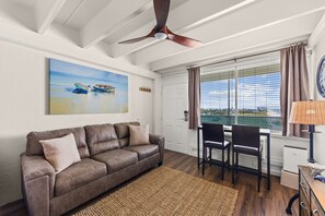 A living room with a brown leather sofa, a dining table with chairs, a ceiling fan, a painting of boats, and a window with curtains. - Beachfront Studio at Fontainebleau | Private Beach Access (Panama City Beach)