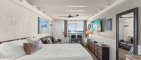 A bedroom with a large bed, a desk, and a chair. There is a window with curtains, a ceiling fan, and artwork on the walls.