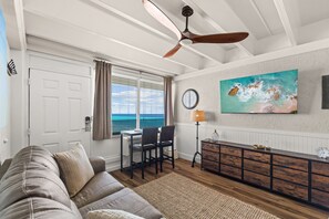 A living room with a sofa, dining table, chairs, a ceiling fan, a painting, and a view of the sea through a window. - Beachfront Studio at Fontainebleau | Private Beach Access (Panama City Beach)