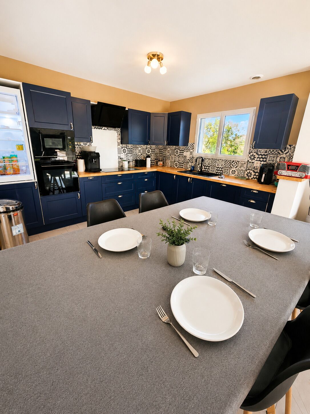 Ett modernt kök med mörkblå skåp, ett grått matbord med plats för fyra personer och ett fönster med utsikt över grönområden.