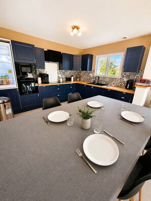 A modern kitchen with dark blue cabinets, a grey dining table set for four, and a window with a view of greenery. - Haven of Peace (Gommenec'h)