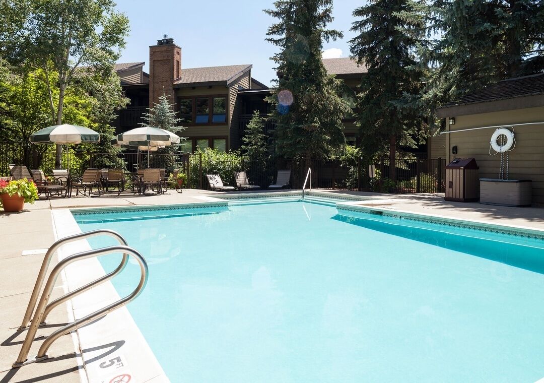 A swimming pool with a lifeguard chair and lounge chairs, surrounded by trees and a building with a brick chimney.