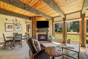 A spacious living room with a fireplace, a flat-screen TV mounted above it, and large windows offering a view of the outdoors. - 2BR Mountain Condo | Walk to Resort, Hot Tubs, Pool (Steamboat Springs)