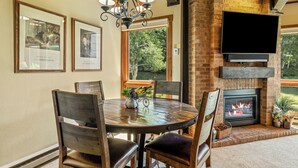 A dining area with a brick fireplace, a flat-screen TV mounted above it, and framed pictures on the wall. - 2BR Mountain Condo | Walk to Resort, Hot Tubs, Pool (Steamboat Springs)