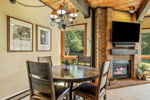 A dining area with a brick fireplace, a flat-screen TV mounted above it, and framed pictures on the wall. - 2BR Mountain Condo | Walk to Resort, Hot Tubs, Pool (Steamboat Springs)