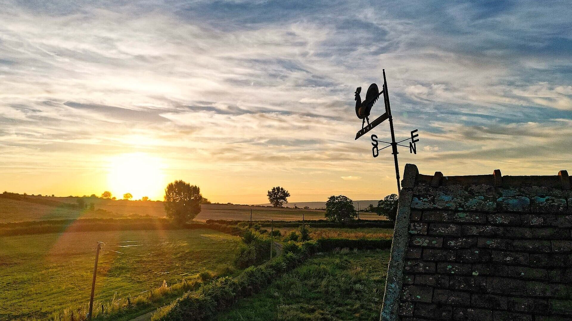Une girouette fixée sur un mur de briques, avec un paysage rural en arrière-plan.