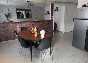 A dining area with a brick wall, a round wooden table, black chairs, a white coffee pot, and a refrigerator.