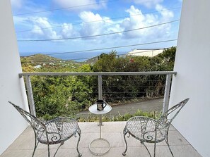 Ein Balkon mit zwei Metallstühlen und einem kleinen runden Tisch, der eine üppige Landschaft mit Hügeln und wolkenlosem Himmel bietet.