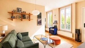 A living room with a green sofa, a glass coffee table, and a wooden shelf with pictures. There is a window with a view of trees. - Cocon cosy et lumineux · 1BDR · Bastille (Paris)