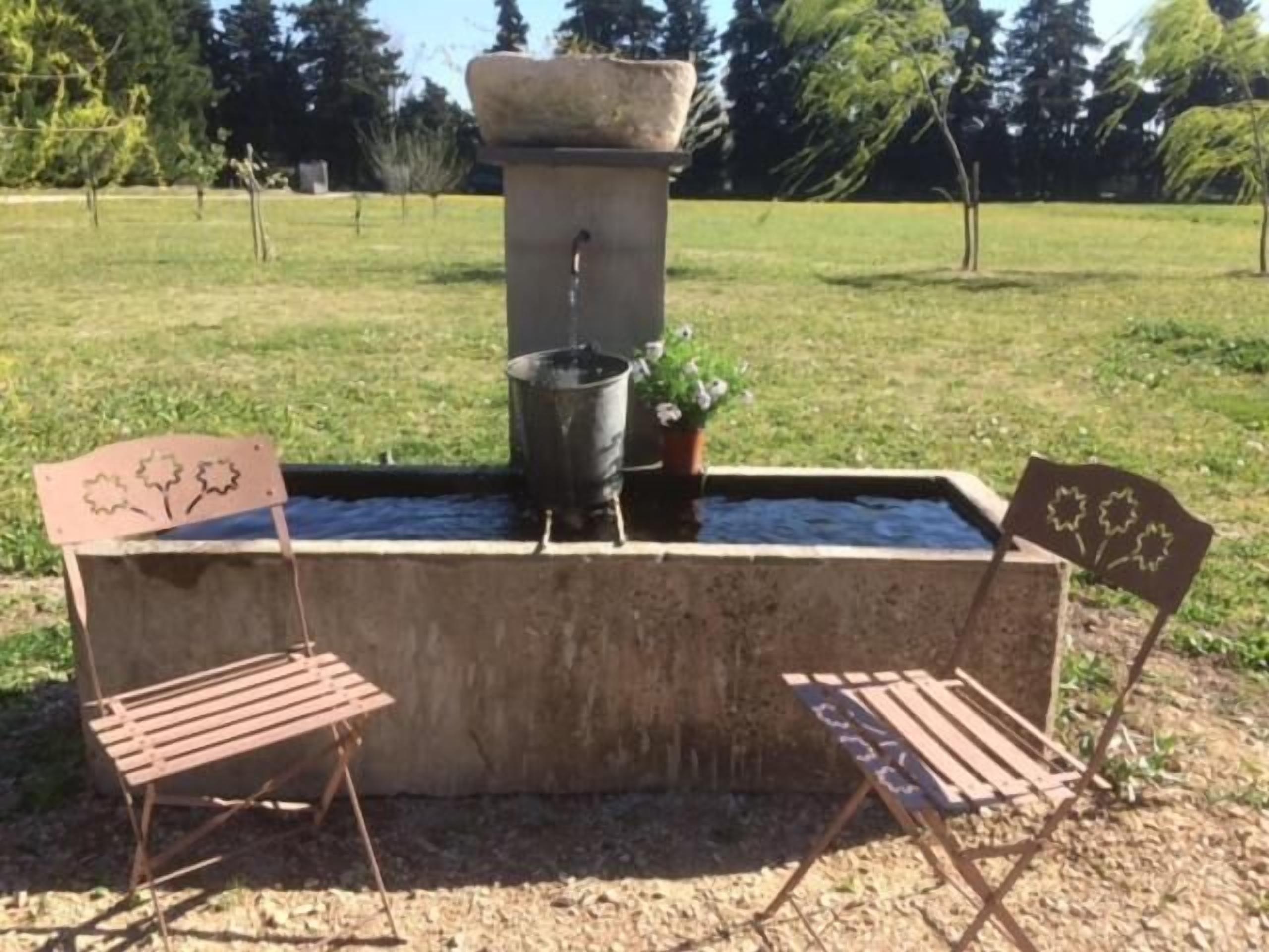 Ein Beton-Wasserspiel mit einer Steinschale und einem Metall-Eimer, umgeben von zwei Metallstühlen mit floralen Verzierungen.