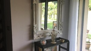 A room with a large ornate mirror on a black console table, a framed picture, and a window with a view of greenery.