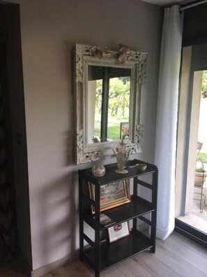 A room with a large ornate mirror on a black console table, a framed picture, and a window with a view of greenery.