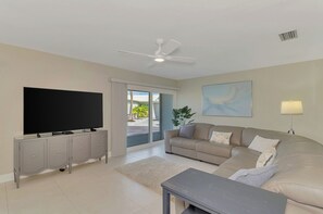 A living room with a flat-screen TV, a ceiling fan, a sofa, a coffee table, and a sliding glass door leading to an outdoor area. - Remodeled Siesta Key Villa – Steps to Private Beach & Village! (Sarasota)