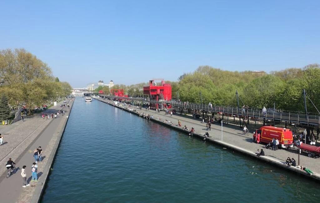 Een gracht met een voetpad, een rode constructie en een brug in de verte.
