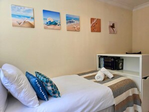 Una cama bien hecha con almohadas, una manta a rayas y una toalla enrollada. Hay cuatro cuadros en la pared, una estantería con libros y una mesita de noche blanca.