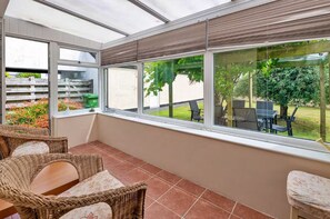 Une véranda meublée de mobilier en osier, dotée de grandes fenêtres et offrant une vue sur un jardin.