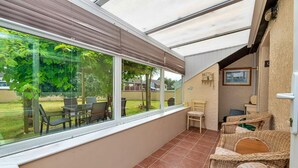 Ein sonnendurchfluteter Raum mit einem großen Fenster, eine Terrasse mit Stühlen und ein kleiner Tisch.
