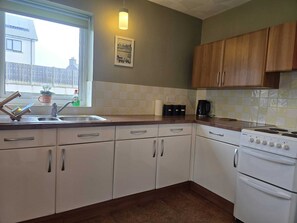 A kitchen with white cabinets, a sink, a stove, and a window with a view of houses. - Peaceful Property North of the island - Pass the Keys (Amlwch)