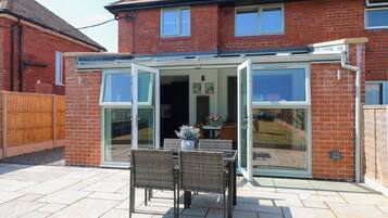 A house with a glass-enclosed patio, a dining table with chairs, and a garden visible through the glass.