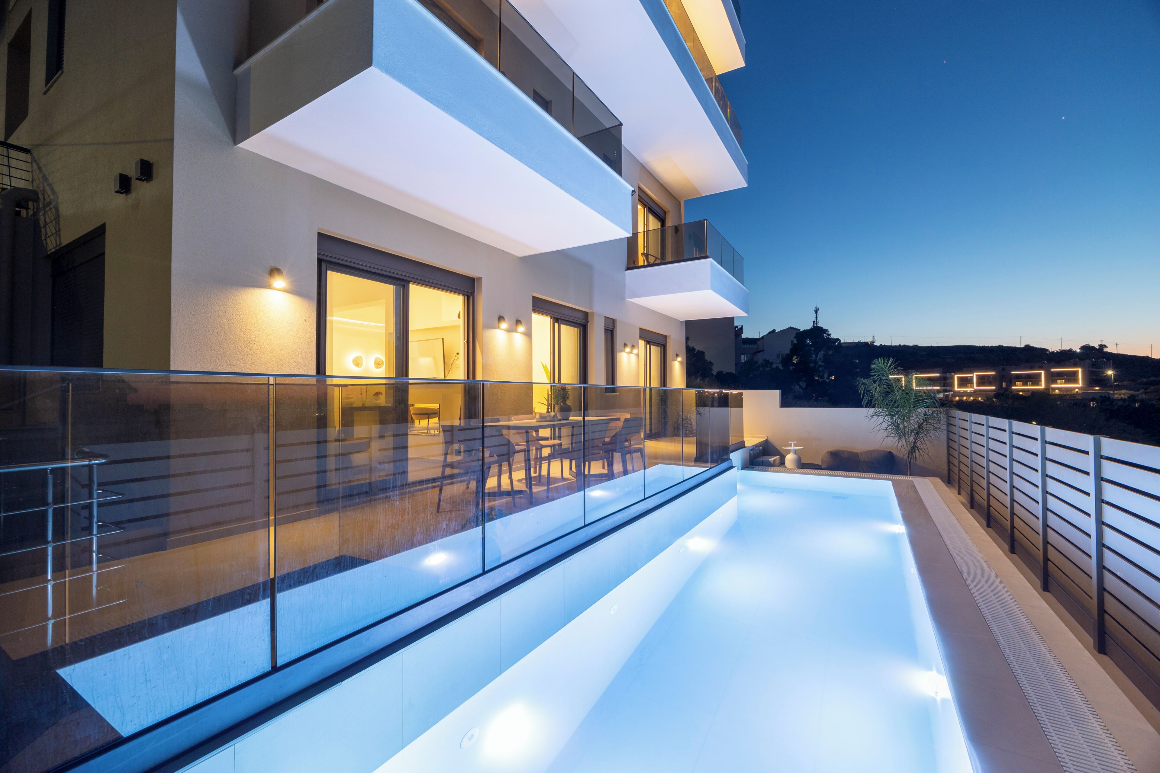 A modern building with a glass balcony, a swimming pool, and a dining area with outdoor furniture.