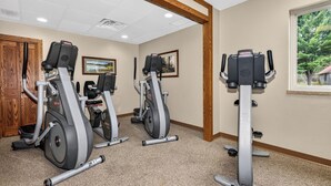 Uma academia com três equipamentos elípticos, uma porta de madeira e uma janela com vista para árvores.