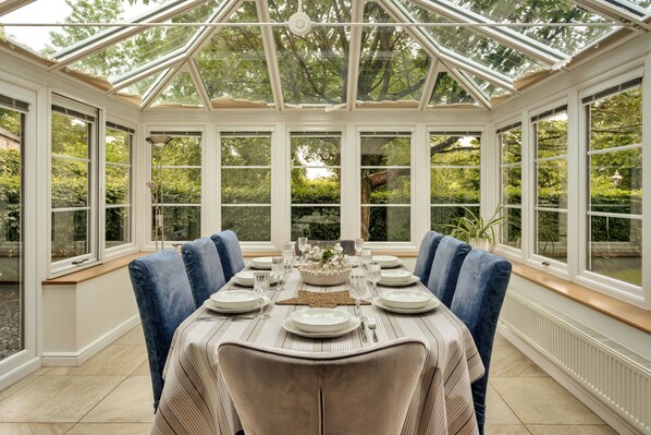 A conservatory with a glass roof, a dining table set for six, and a view of greenery outside.