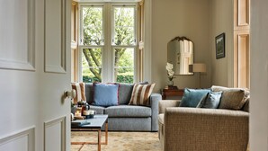 Ein Wohnzimmer mit Sofa, Sessel, Couchtisch und Blick auf Grün durch große Fenster.