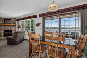 A dining area with a stone fireplace, wooden chairs, and a view of a lake through large windows.