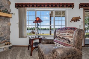 A room with a stone wall, a window with a view of a lake, a lamp, a leather armchair, and a bear wall decoration.