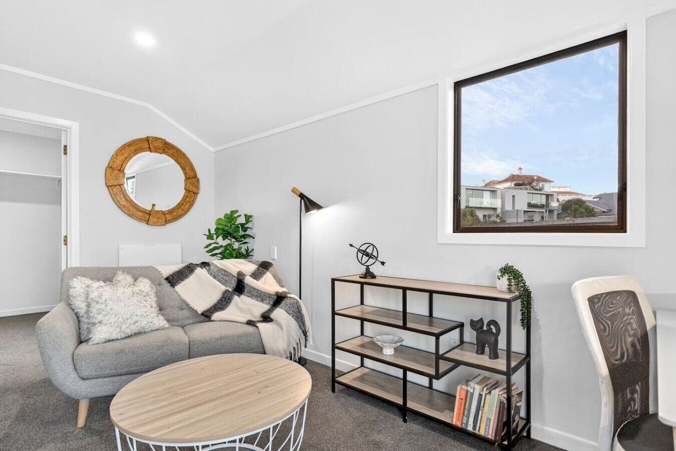 Uma sala de estar moderna com um sofá cinzento, um espelho redondo, uma estante de livros e vista para um edifício através de uma janela.