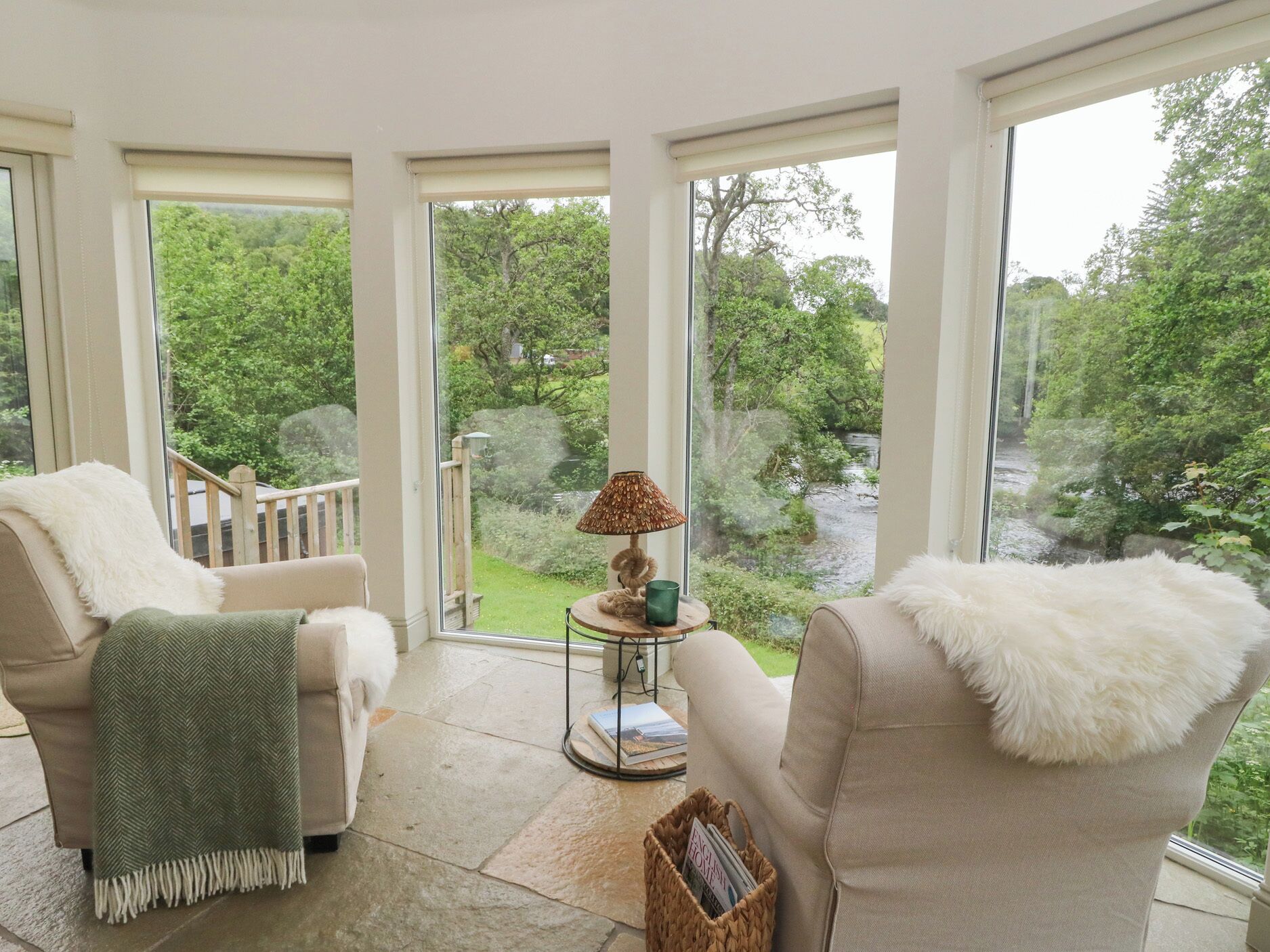 Una habitación con dos sillones color beige, una mesita redonda con lámpara y vistas a un paisaje verde a través de amplios ventanales.