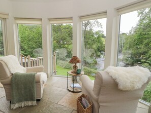 A room with two beige armchairs, a small round table with a lamp, and a view of a green landscape through large windows. - Glen Lochay Lodge (Killin)