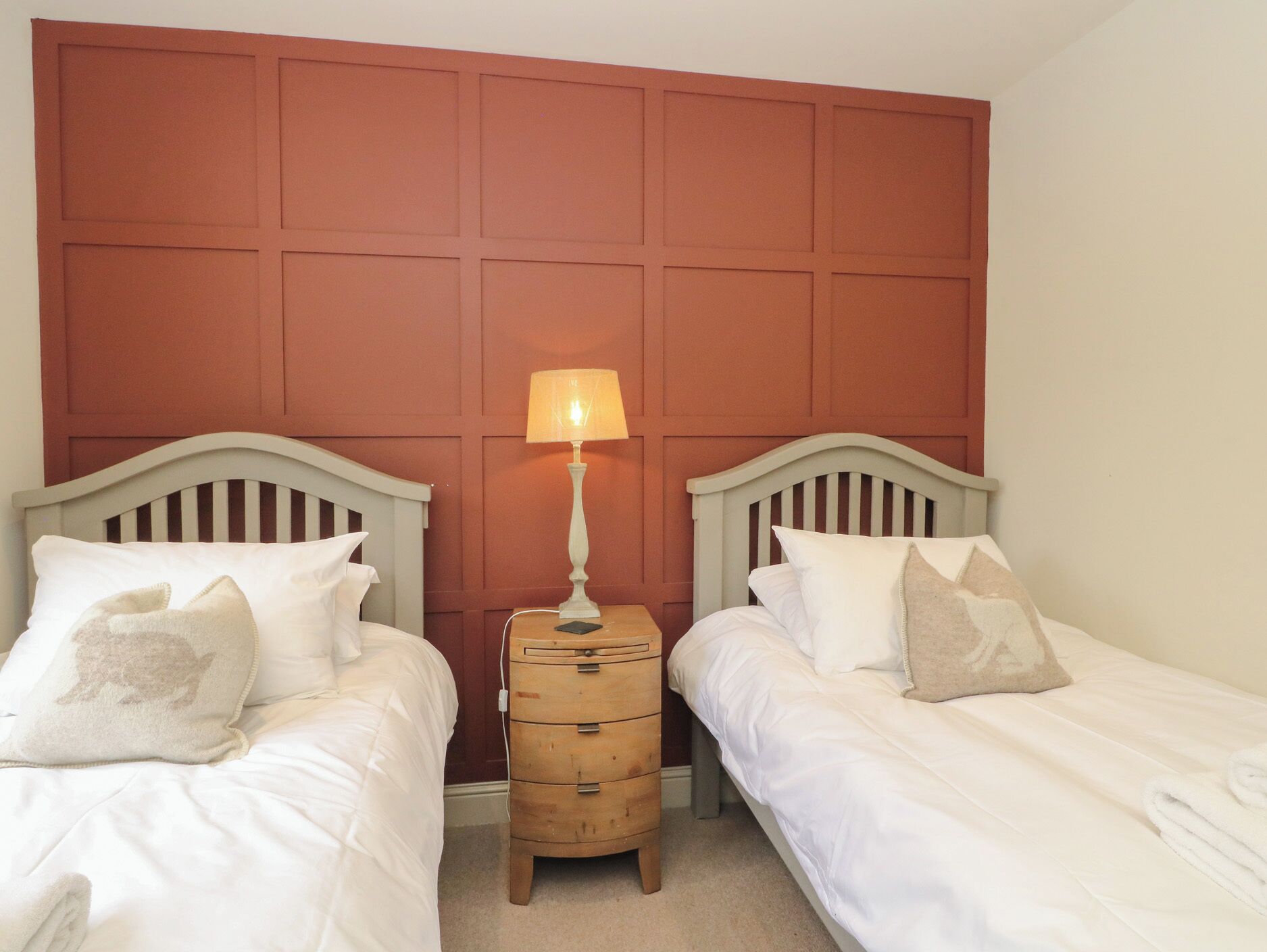Um quarto com duas camas de solteiro, uma mesa de cabeceira com luminária e um aparador de madeira.
