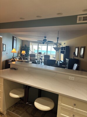 A kitchen with a marble countertop, two stools, a ceiling fan, and a view of the ocean. - Gulf-front paradise in Springtime on lovely Longboat Key 2/2 -2 pools,pickleball (Longboat Key)