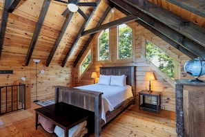 Ein Schlafzimmer einer Holzhütte mit einem Bett, Nachttischlampen, einem kleinen Tisch und einem Fenster mit Blick auf Bäume.