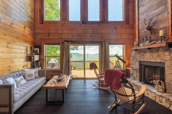 L’intérieur d’une cabane en rondins, avec une cheminée, un canapé, un fauteuil à bascule et une vue sur l’extérieur.
