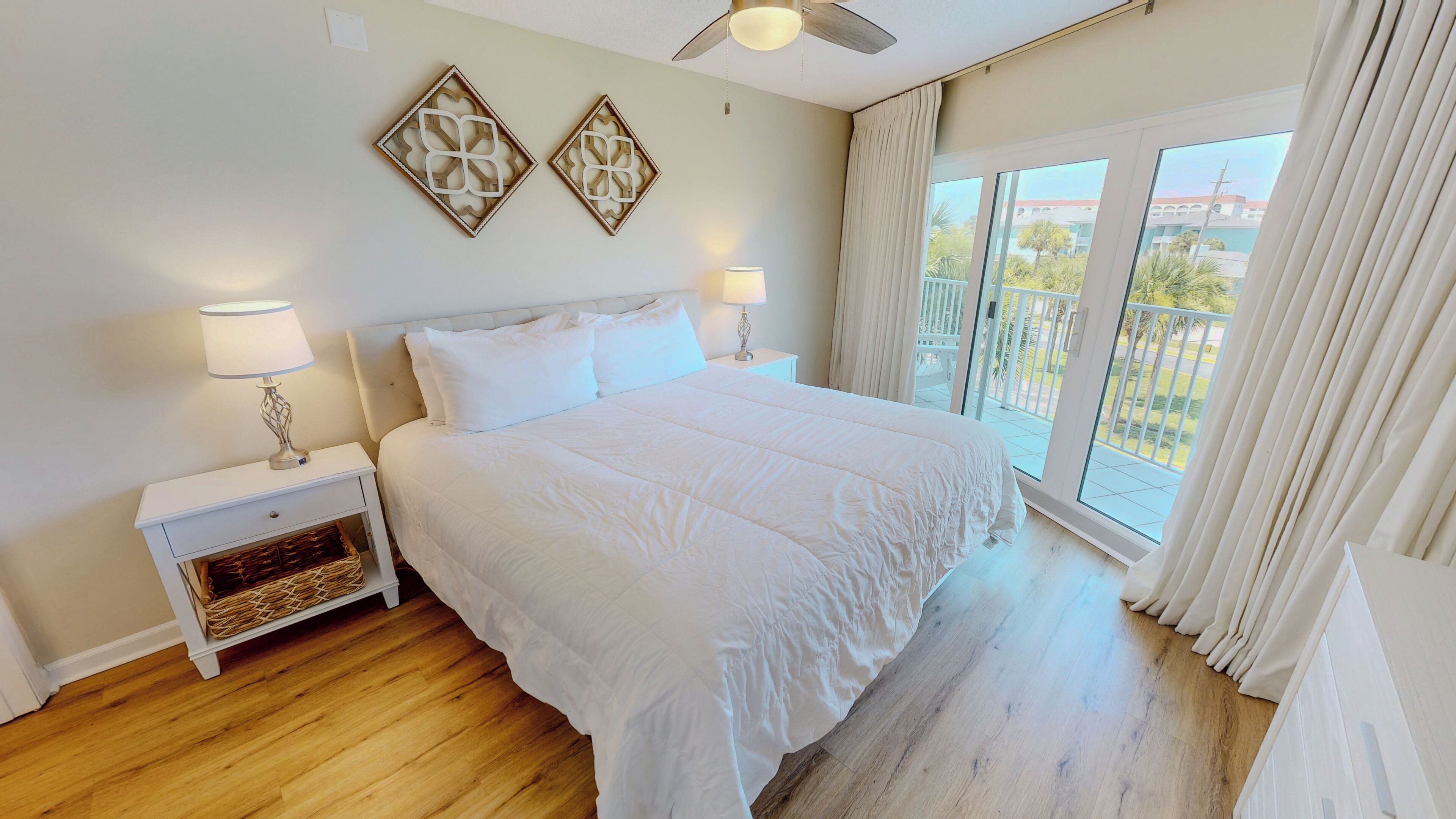 Une chambre avec un grand lit, deux tables de chevet, un ventilateur de plafond et un balcon avec des rideaux.