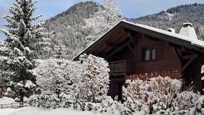 Eine Holzhütte mit geneigtem Dach, umgeben von schneebedeckten Bäumen und einer Bergkulisse.