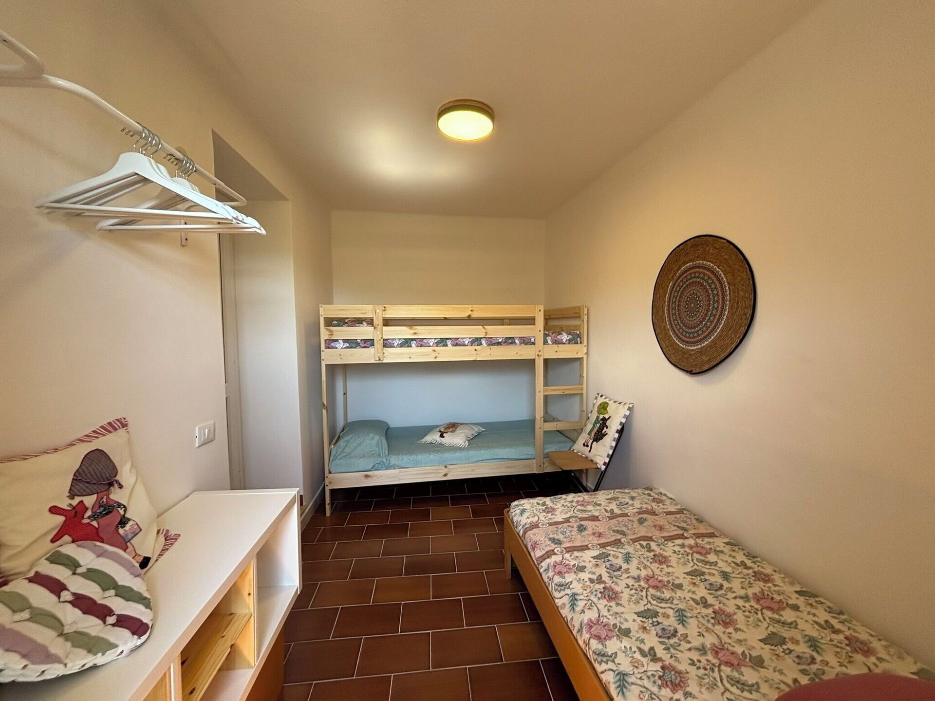 Quarto pequeno e compacto com uma beliche, uma cama de solteiro, uma prateleira branca com um almofada e um chapéu fixado na parede.