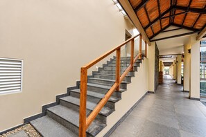 Una escalera con barandillas de madera y peldaños de piedra, que conduce a un pasillo con techo alto y ventanas amplias.