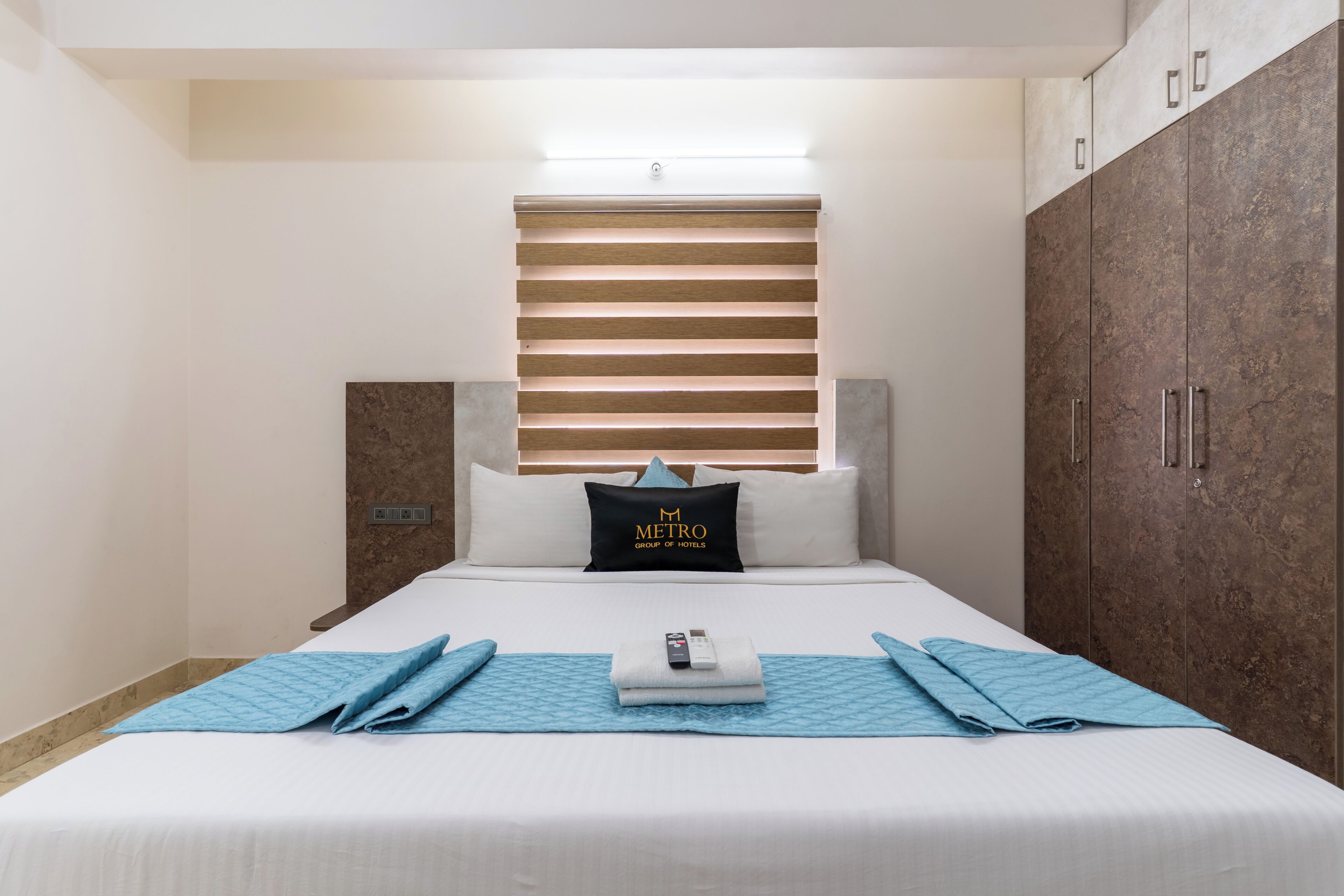 Habitación de hotel con cama, cabecera con listones horizontales, almohada con el texto 'METRO GROUP OF HOTELS', mesita de noche y armario.