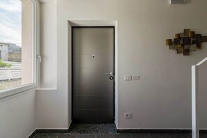 A modern interior with a closed door, a wall-mounted light fixture, and a window with a view of buildings. - KeyRoom • Patio & Parking near Lake Como (Como)