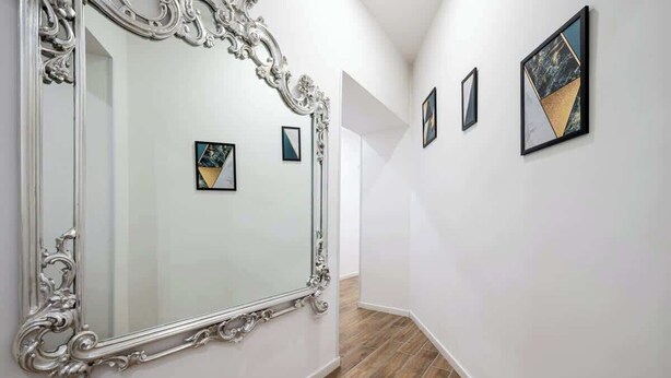 Een gang met een grote, sierlijke spiegel, een houten vloer en kunstwerken in lijsten aan de wanden.
