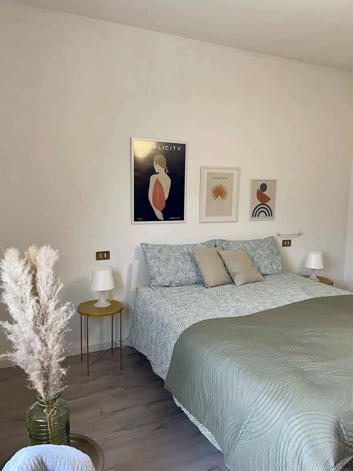 Una camera da letto con un letto, due comodini, una lampada, un vaso con piume di pampa e tre elementi decorativi a parete.